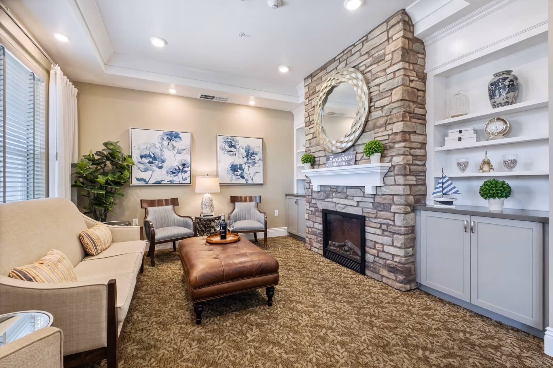 Cozy living room with a stone fireplace topped by a round mirror, built-in shelves, a sofa, chairs and a leather ottoman.