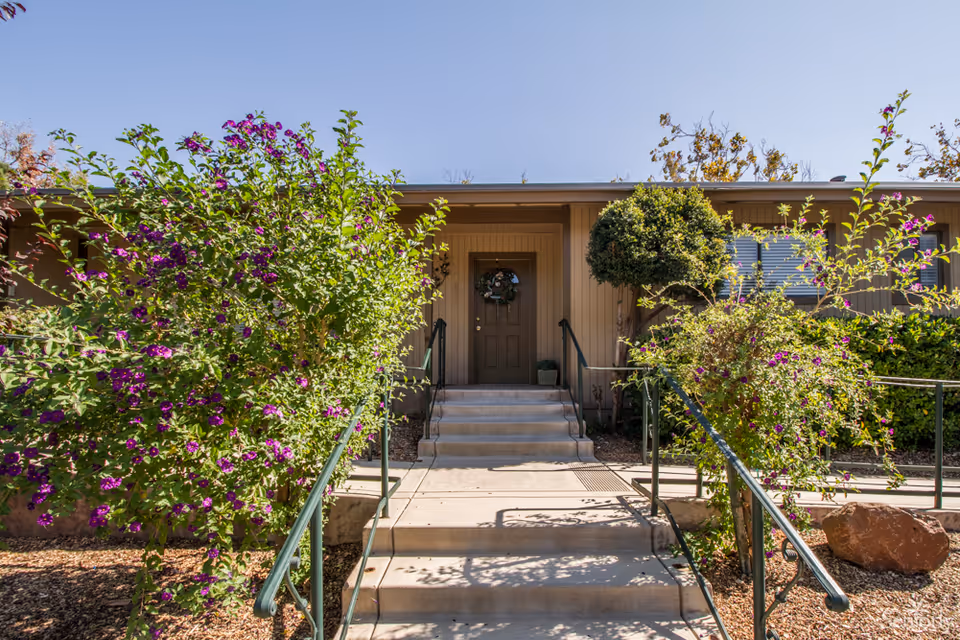 Entrance to a single-story building with a brown door decorated with a wreath. There are concrete steps and a ramp with green handrails leading up to the door. On either side of the walkway, there are bushes with purple flowers and a small tree. The sky is clear and blue.