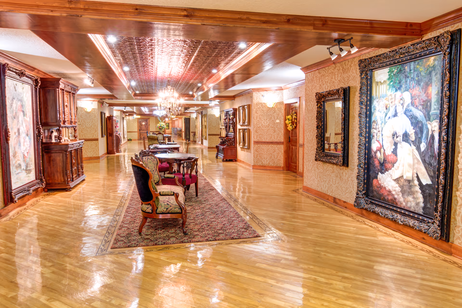 Ornate polished wood-floored corridor with upholstered chairs, small tables, chandeliers, and large framed paintings on the walls.