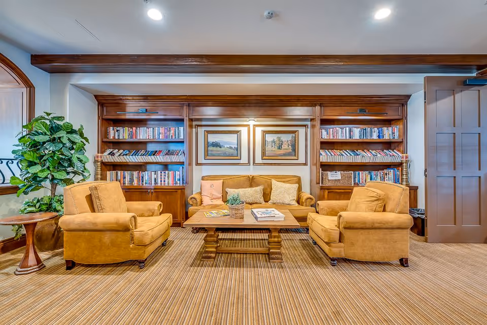 A cozy sitting area in a senior living facility with two mustard yellow armchairs and a matching sofa arranged around a wooden coffee table. Behind the sofa, there are built-in wooden bookshelves filled with books and two framed landscape paintings on the wall. A potted plant and a small round wooden side table are placed to the left of the armchairs. The room has a striped carpet and recessed ceiling lights.