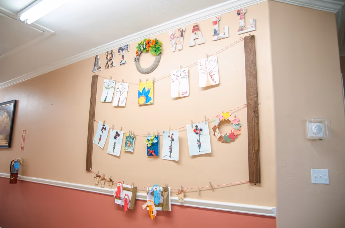 A wall display in a senior living facility with the words 'ART WALL' at the top. Below the letters, several pieces of colorful artwork and crafts are hung on strings with clothespins. The wall is painted beige with a white chair rail and a reddish lower section. A fire extinguisher and a framed picture are visible on the left side of the image.