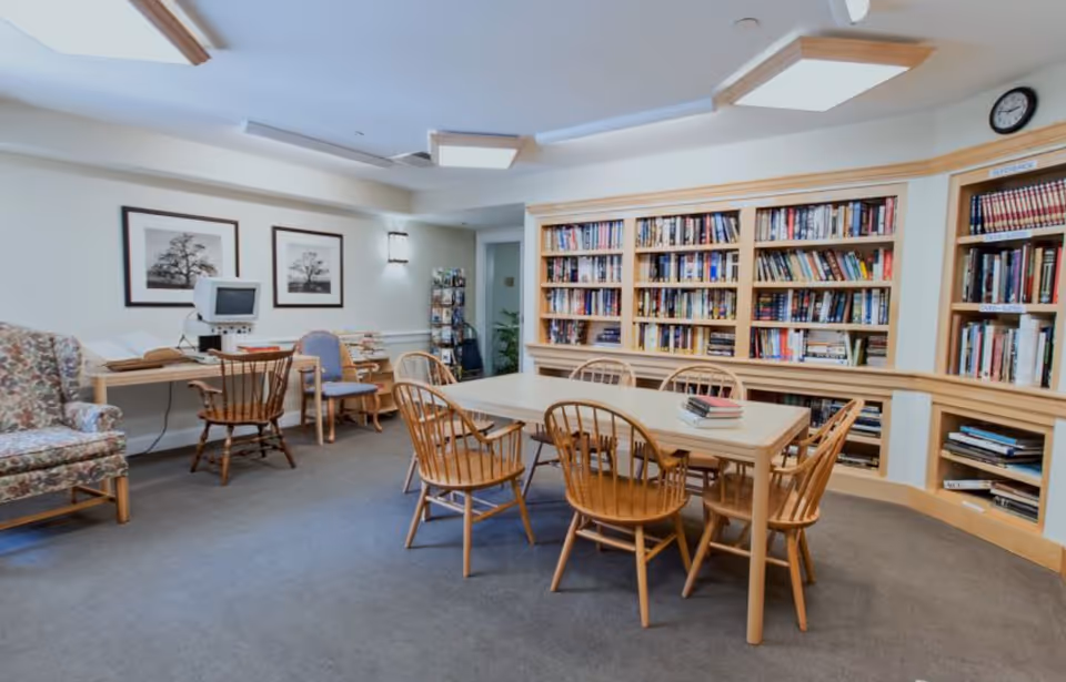 A community library/reading room with a central table surrounded by wooden chairs and bookshelves lining the wall.