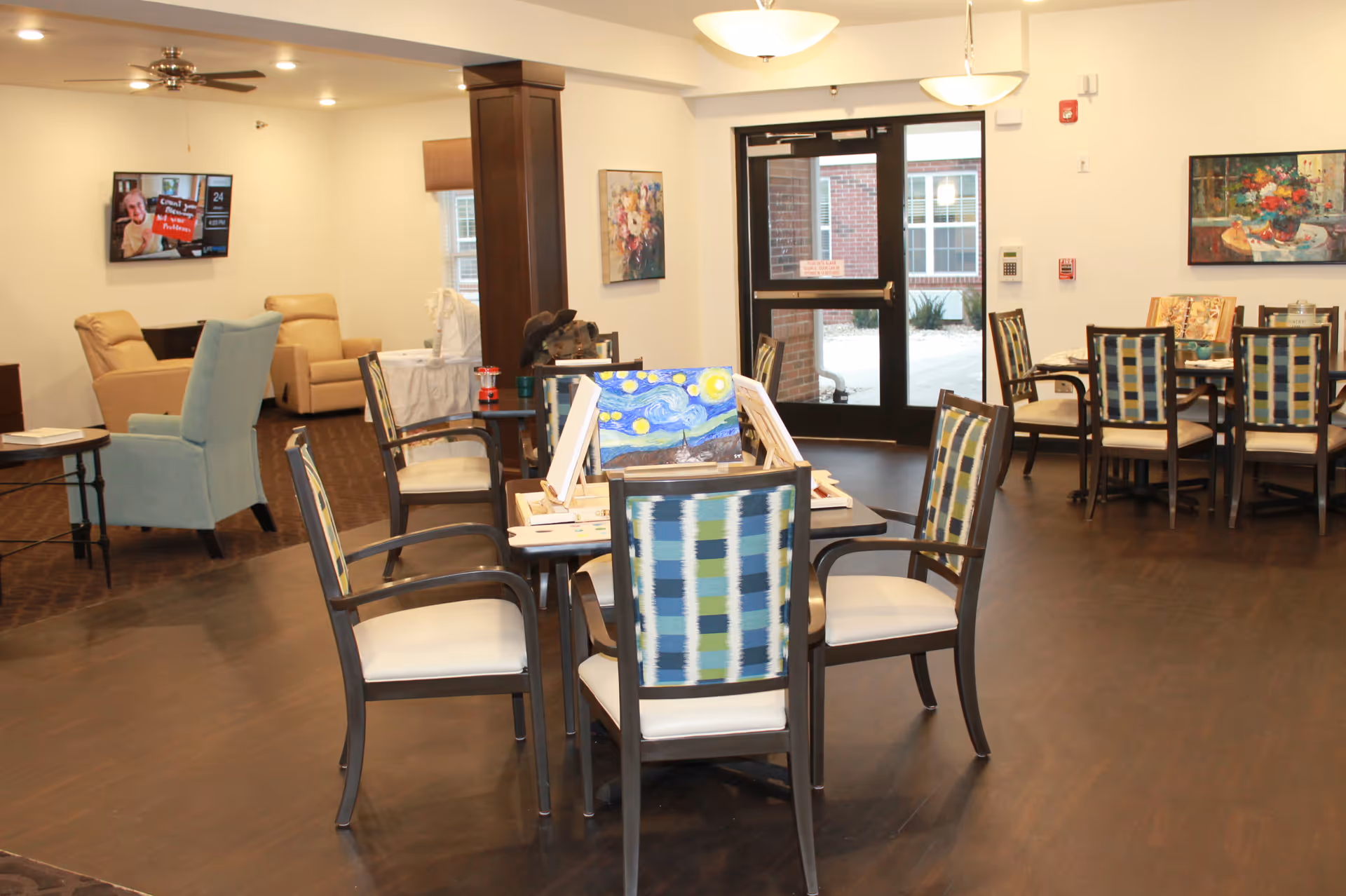 A spacious common area in a senior living facility with multiple tables and chairs arranged for activities. One table has painting supplies and a canvas with a painting resembling Van Gogh's Starry Night. In the background, there are comfortable armchairs, a wall-mounted TV, and large glass doors leading outside. The room is well-lit with ceiling lights and decorated with colorful paintings on the walls.