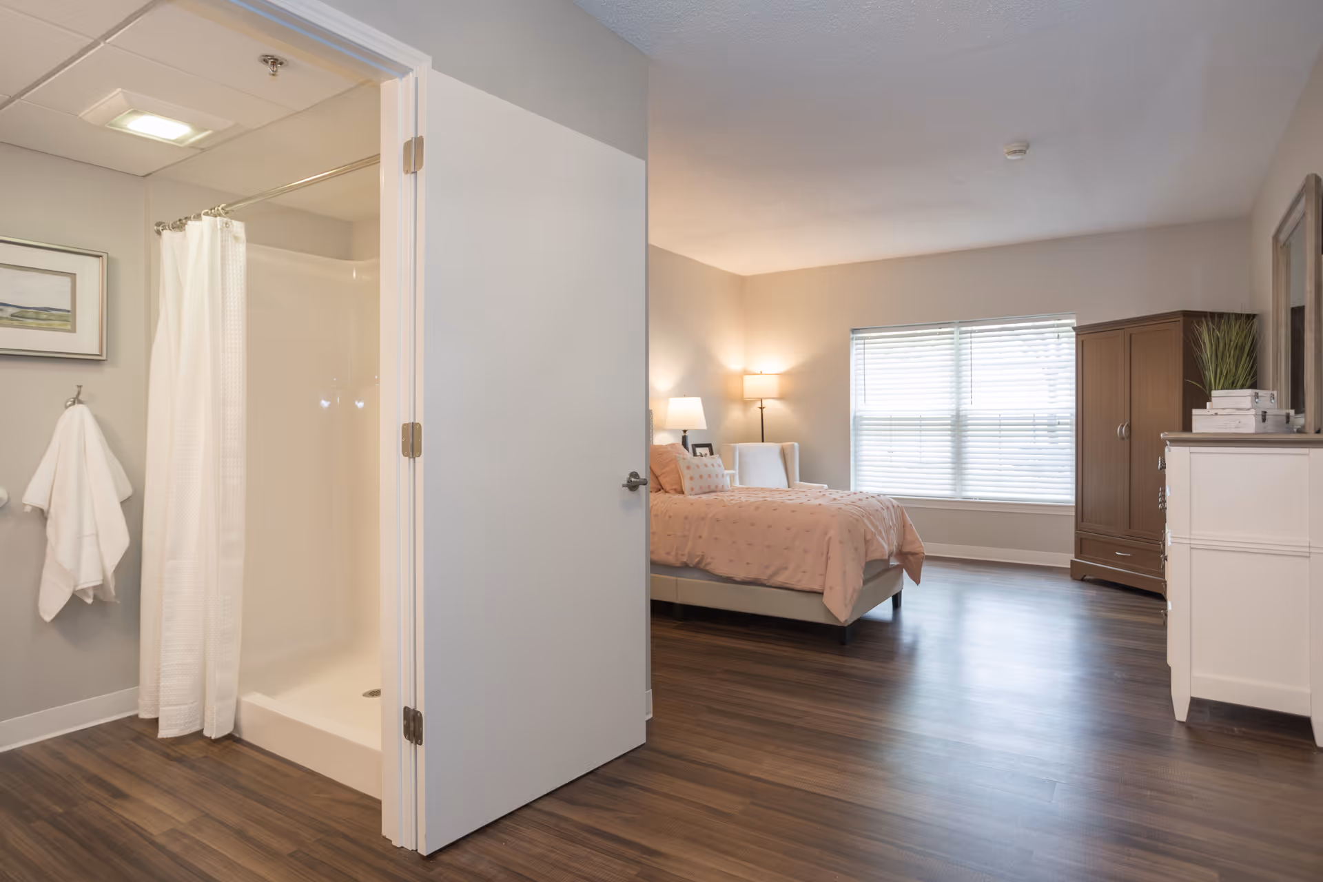 A spacious senior living bedroom with a bed covered in a light pink comforter, a white armchair, a wooden wardrobe, and a white dresser. The room has large windows with blinds letting in natural light. To the left, there is an open door revealing a bathroom with a shower and a white towel hanging on a hook.
