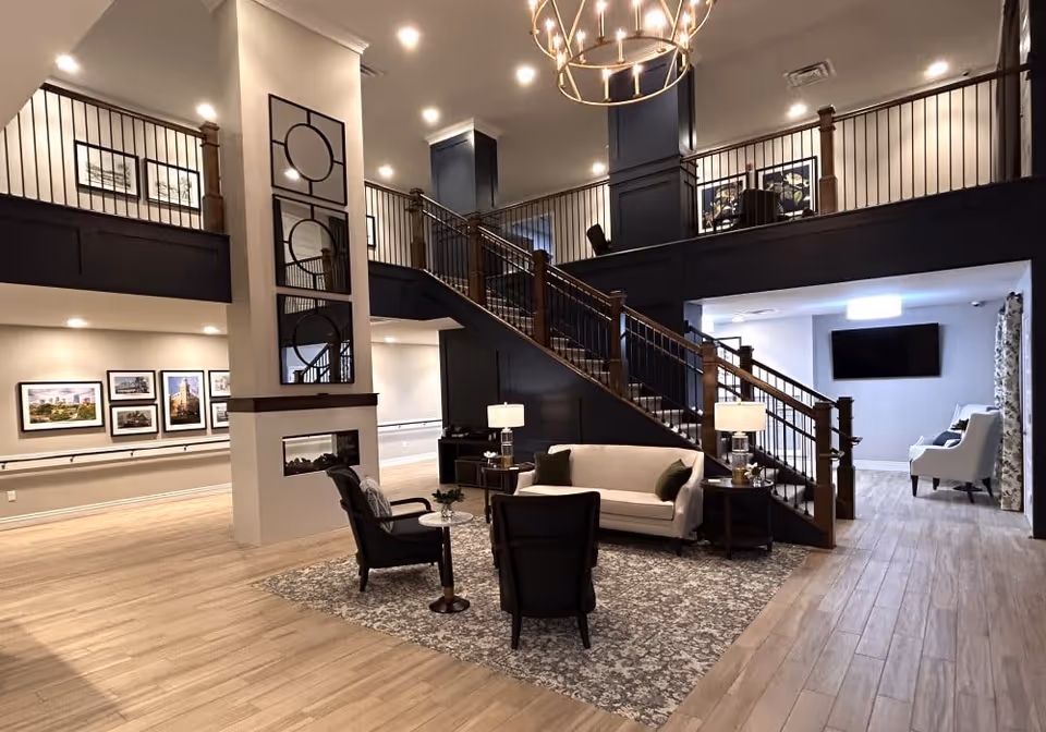 A spacious and modern common area inside a senior living facility featuring a staircase with wooden railings leading to an upper level. The seating area includes a white sofa, two black armchairs, a small round table, and two side tables with lamps, all arranged on a patterned rug. The walls are decorated with framed pictures and mirrors, and there is a flat-screen TV mounted on the wall in the background. The flooring is light wood, and the space is well-lit with recessed lighting and a chandelier.