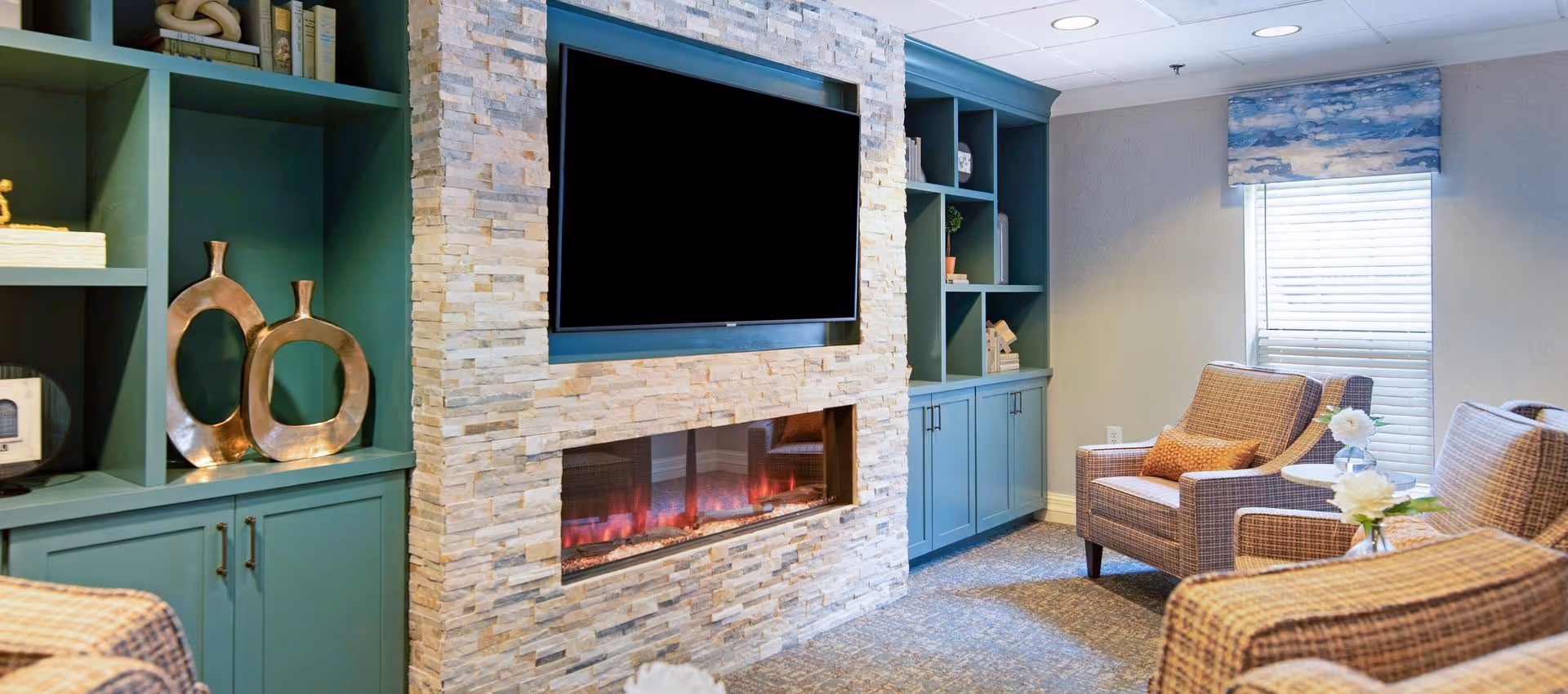 Cozy communal lounge with cushioned armchairs, teal built-in shelving, a stone fireplace and a wall-mounted TV.