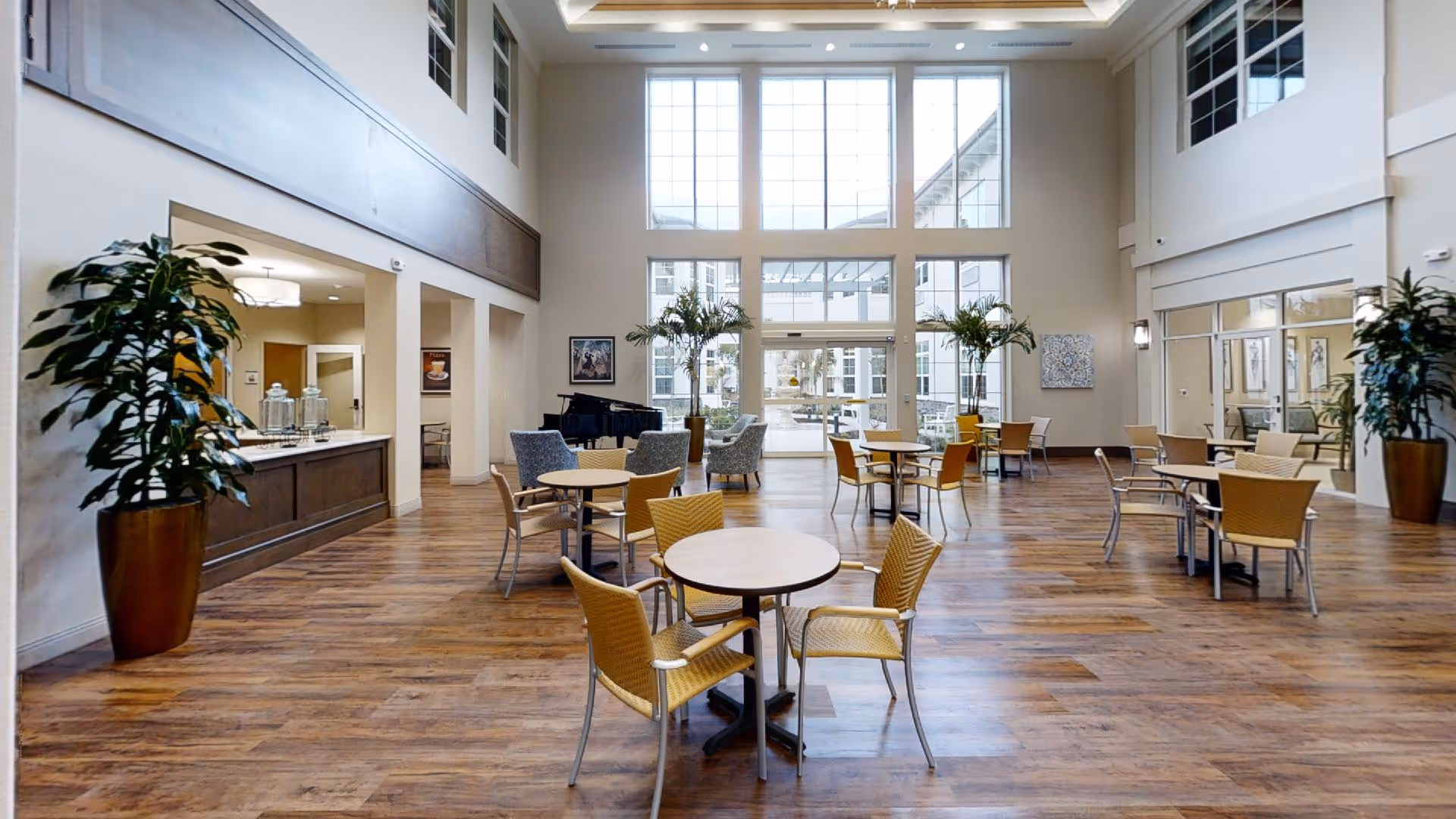 A spacious, well-lit common area with high ceilings and large windows letting in natural light. The room features several round tables with yellow chairs, a few armchairs near the back, and large potted plants. There is a black grand piano against the far wall and a counter area on the left side. The flooring is wood, and the overall decor is modern and inviting.