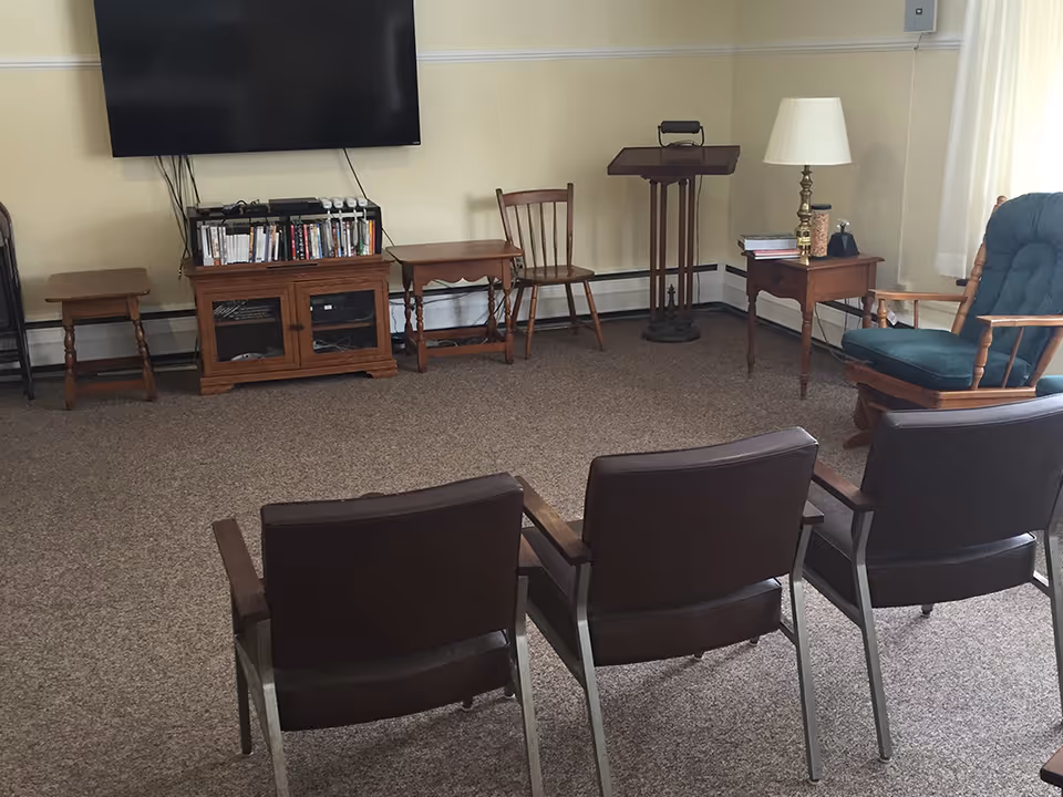 A small sitting area with three brown chairs in the foreground facing a wall-mounted flat screen TV. Below the TV is a wooden cabinet with shelves holding DVDs and electronic devices. There are additional wooden chairs and small tables around the room, including a green cushioned rocking chair and a side table with a lamp. The room has beige walls and carpeted floor.