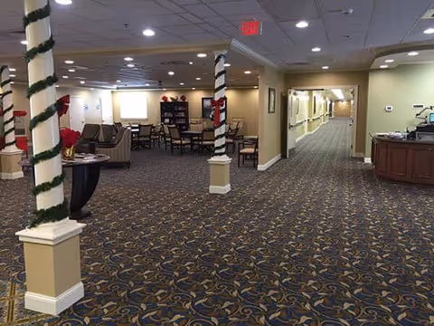 Interior view of a senior living facility common area with carpeted floor, decorated columns wrapped with garland and red bows, a seating area with chairs and tables, and a long hallway leading to other rooms.