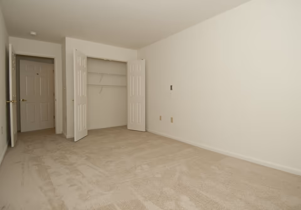 Empty room with beige carpet, white walls, an open closet with double doors, and a closed door leading to another room.
