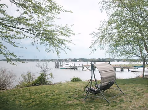 A peaceful outdoor scene showing a grassy area with a single cushioned swing chair facing a body of water. In the background, there are several boats docked at a marina, with trees framing the view on both sides.