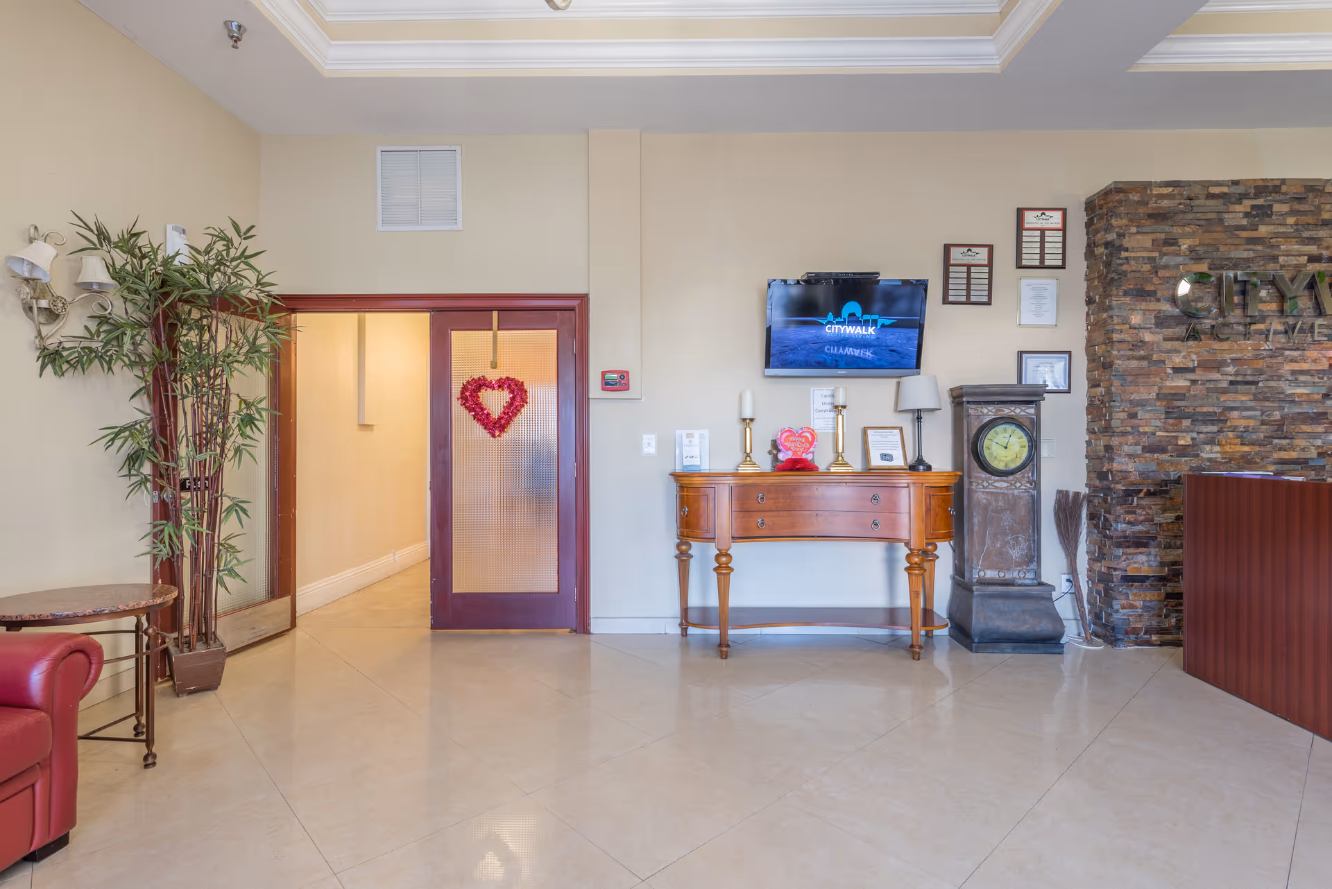 Interior view of a senior living facility lobby area with a wooden console table holding decorative items, a wall-mounted TV displaying 'Citywalk Clearwater', a tall grandfather clock, a stone accent wall with partial signage, a red leather chair, a potted plant, and a door with a red heart decoration hanging on it.