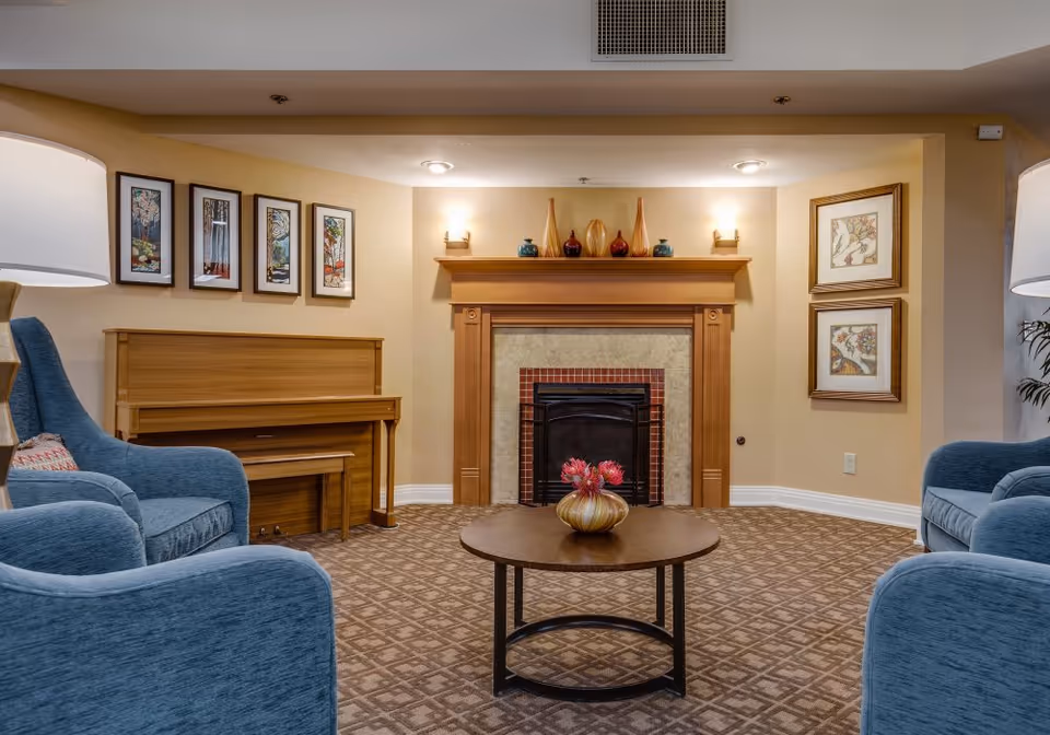 A cozy living room area with a wooden fireplace in the center, decorated with vases on the mantel. There are two blue upholstered armchairs facing a round wooden coffee table with a decorative vase of flowers. A wooden piano with a matching bench is against the left wall, which is adorned with four framed pictures. The right wall has two framed artworks. The room has warm lighting and patterned carpet flooring.