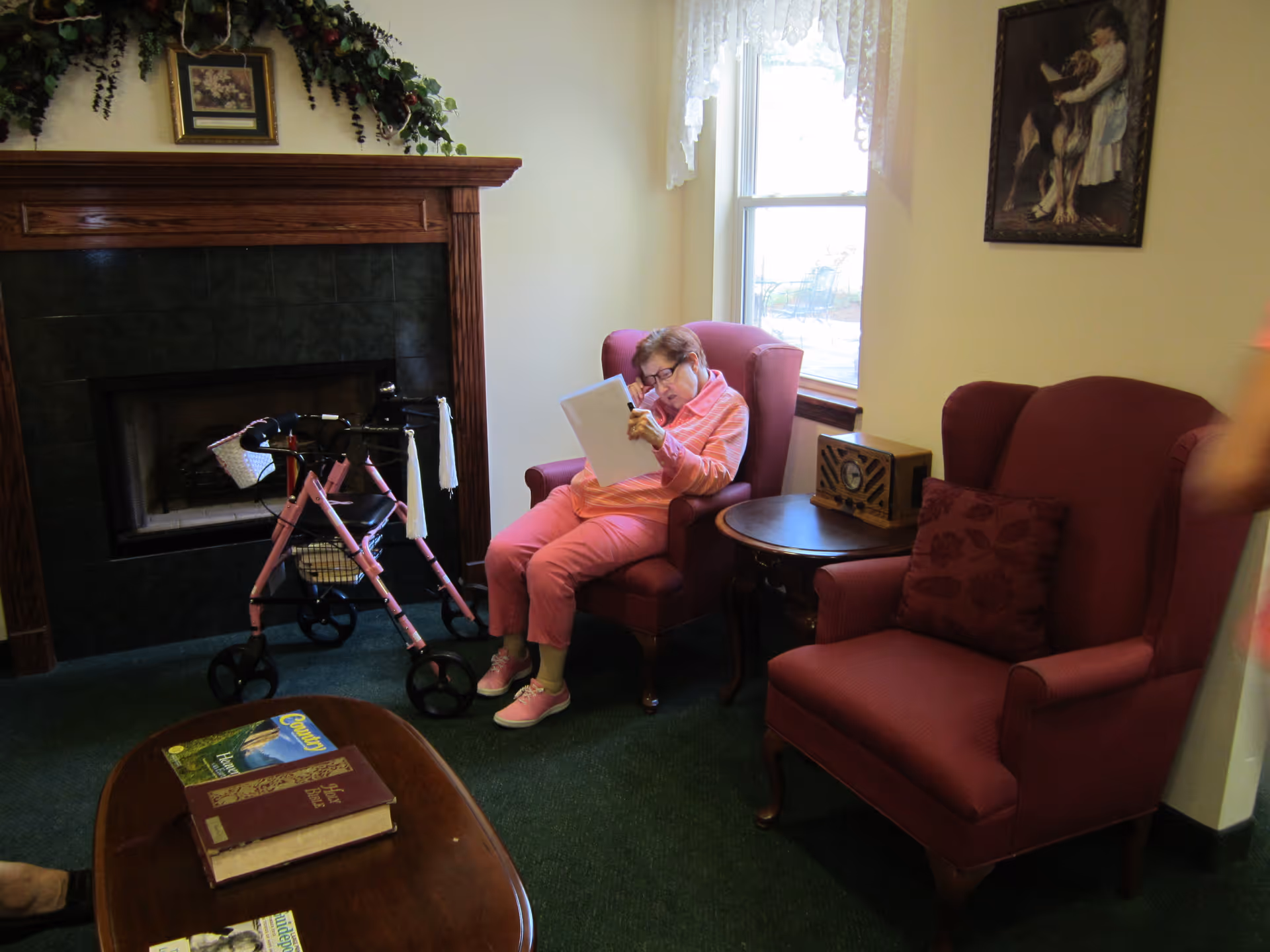 An elderly woman in pink sits reading in an armchair next to a walker and a fireplace in a cozy common room.