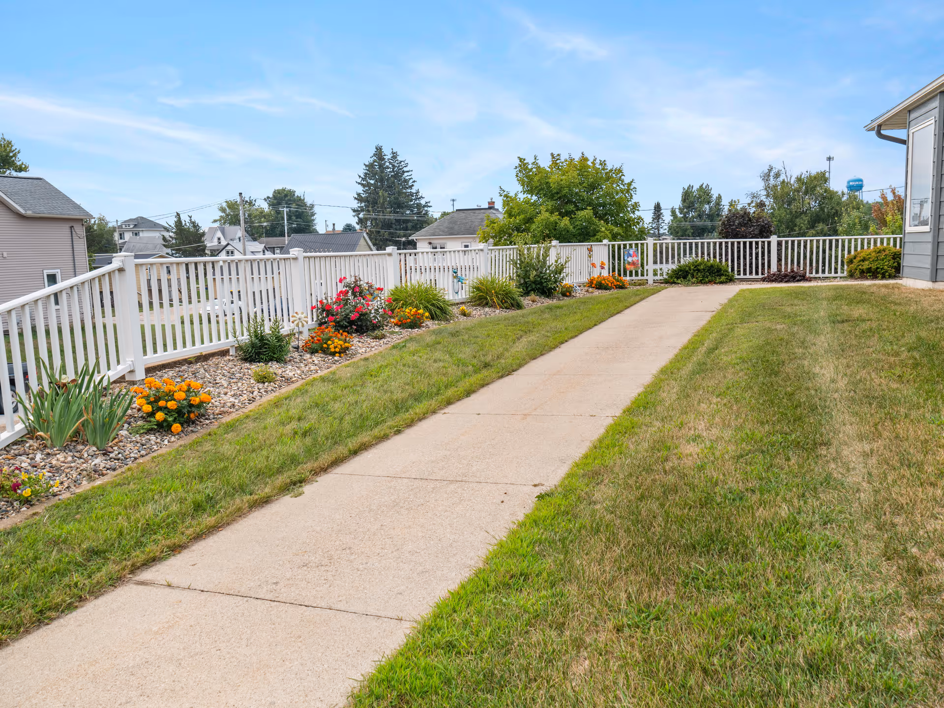A paved walkway bordered by green grass and colorful flower beds with a white fence along the left side. Residential houses and trees are visible in the background under a blue sky with light clouds.