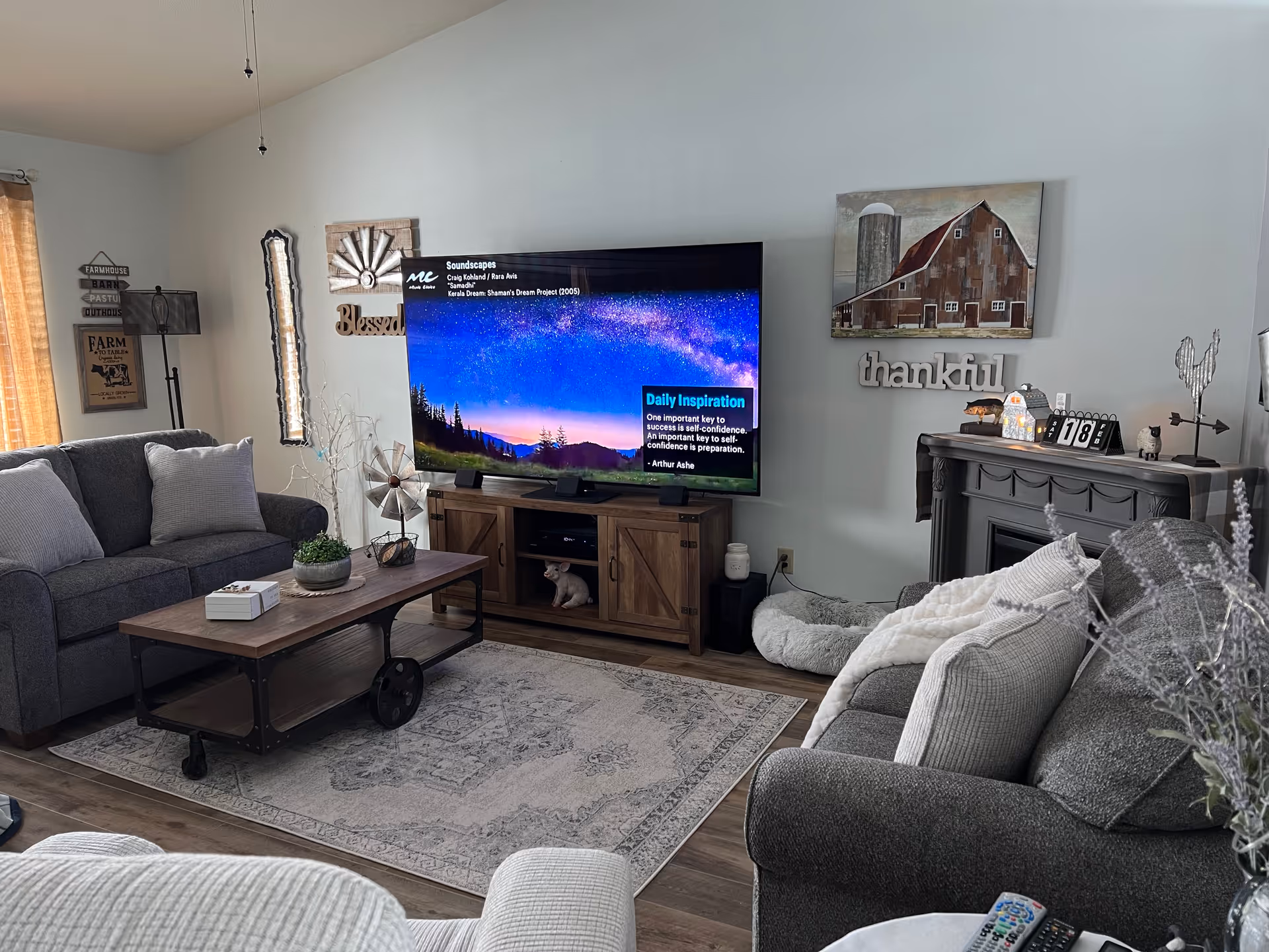 A cozy living room with two gray sofas adorned with light-colored pillows, a wooden coffee table with a small plant and a box on it, a large flat-screen TV displaying a scenic night sky and daily inspiration quote, and various rustic wall decorations including signs that say 'Blessed' and 'thankful'. There is a fireplace with decorative items on the mantel and a dog bed on the floor nearby.