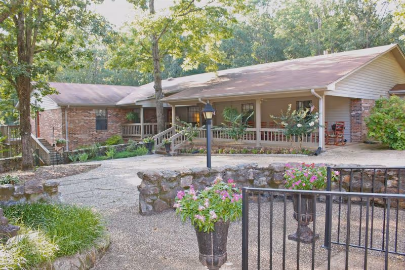 Exterior view of a single-story brick and siding building with a covered porch, surrounded by trees and greenery. There is a stone wall and a paved walkway leading to the entrance, with flower pots containing pink flowers near a black metal fence.