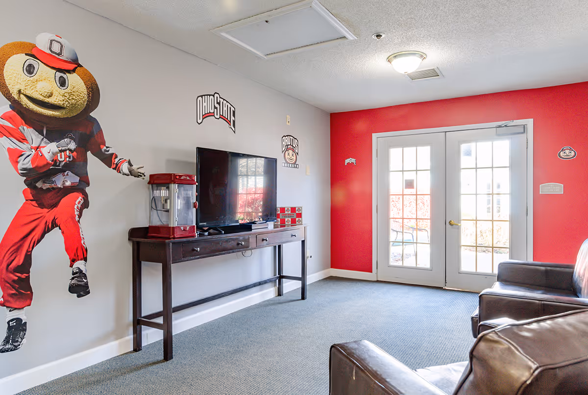 A bright communal living room with a TV on a console, Ohio State mascot wall decal, a popcorn machine, leather chairs, and double French doors set into a red accent wall.