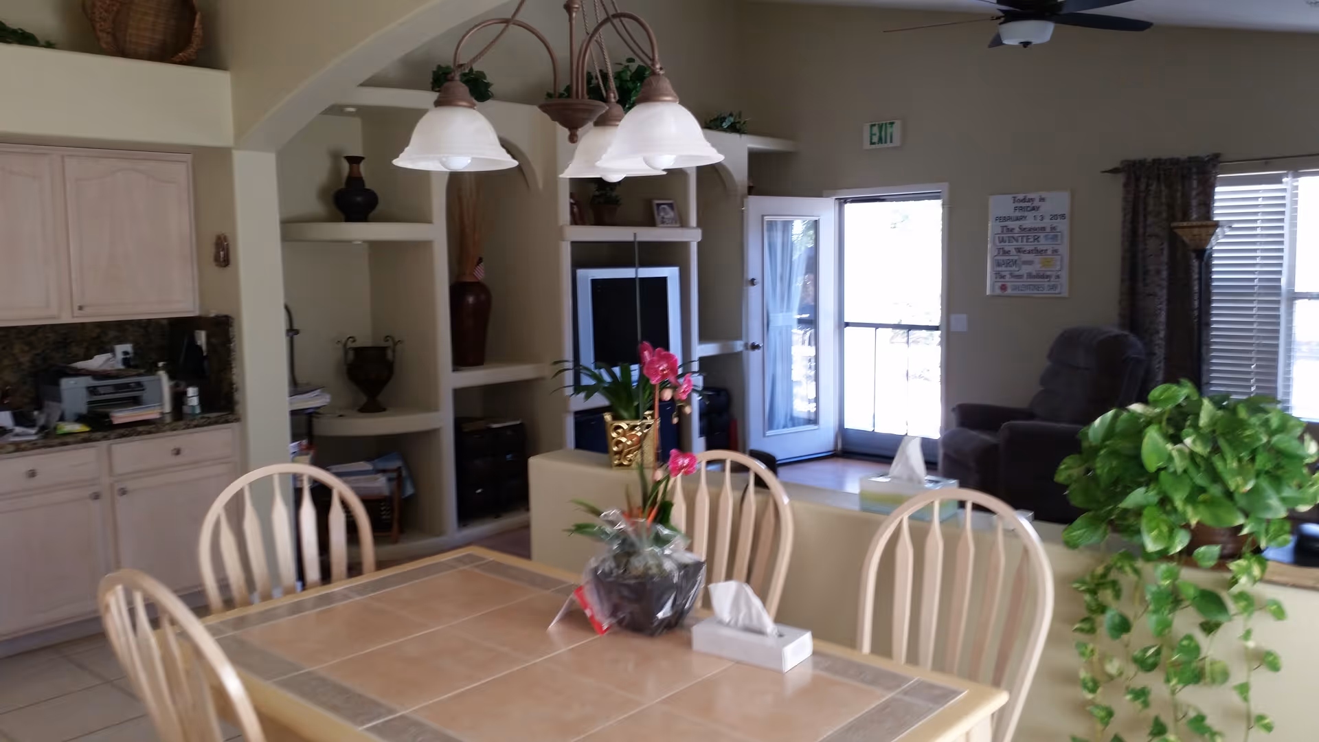 Interior view of a living and dining area in an assisted living facility. The foreground shows a wooden dining table with a tiled surface, surrounded by wooden chairs. On the table, there is a potted plant with pink flowers and a box of tissues. In the background, there is a living room area with a dark recliner chair, a TV on a built-in shelf unit, decorative vases, and a glass door leading outside. A ceiling fan and light fixture hang from the ceiling, and there are windows with blinds and curtains. A green leafy plant is visible on the right side near the dining area.