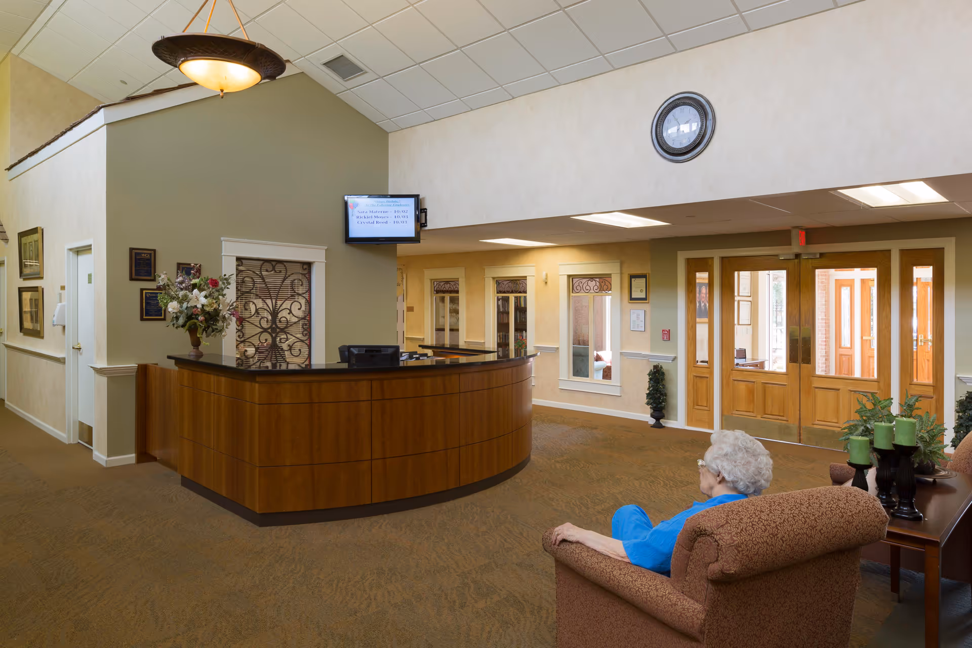 An elderly woman with white hair wearing a blue shirt sits in a patterned armchair in a spacious reception area. The room features a wooden reception desk with a flower arrangement, a wall-mounted TV displaying birthday announcements, a large clock on the wall, and double wooden doors leading to another area. The walls are painted in neutral tones with decorative trim and framed pictures.