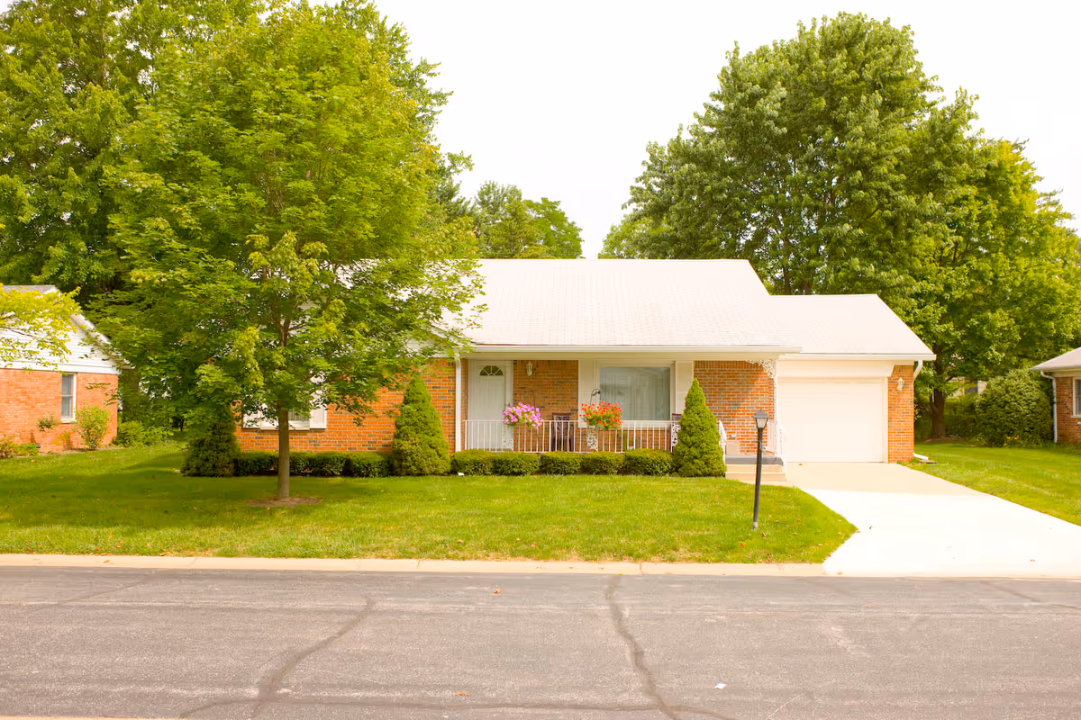 Single-story brick house with a white roof and garage, surrounded by green trees and a well-maintained lawn, with a paved driveway and a street in the foreground.