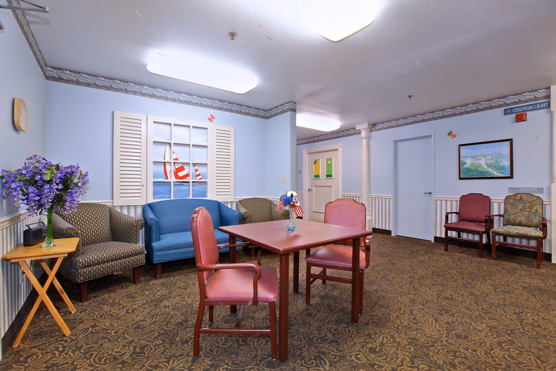 A cozy common area in a senior living facility featuring a square wooden table with four red cushioned chairs around it. Behind the table, there is a blue loveseat and two patterned armchairs. A small wooden side table with a vase of purple flowers is on the left. The walls are light blue with white wainscoting and decorative trim near the ceiling. A window with white shutters shows a painted scene of red and white sailboats on the water. On the right wall, there is a framed landscape painting and two additional chairs, one red and one patterned. The carpet has a brown and gold ornate pattern, and the ceiling has fluorescent lighting.