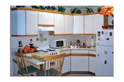 Compact kitchen with white cabinets and appliances, a small breakfast table with two chairs, and decorative items on the countertops and above the cabinets.