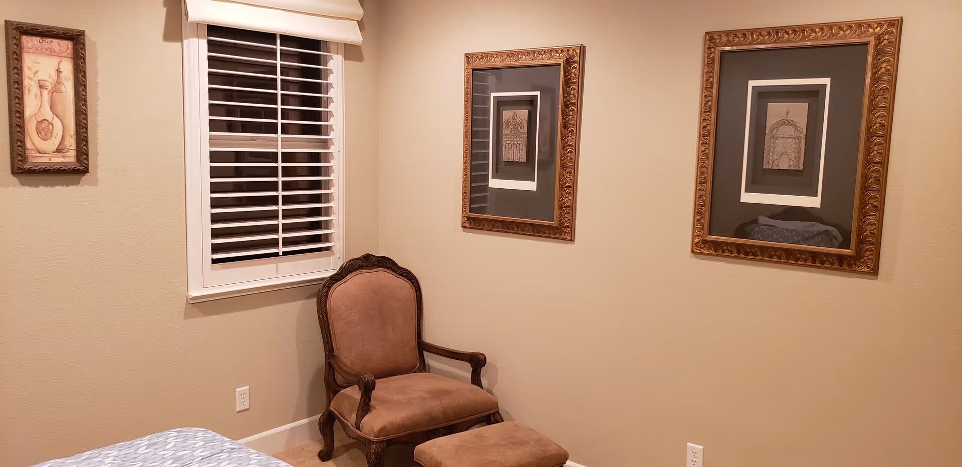 A cozy corner of a bedroom with a cushioned armchair and matching ottoman in front of a beige wall. The wall features two framed pictures with ornate gold frames and a window with white blinds and a beige valance. Part of a bed with a patterned blue and white bedspread is visible in the foreground.
