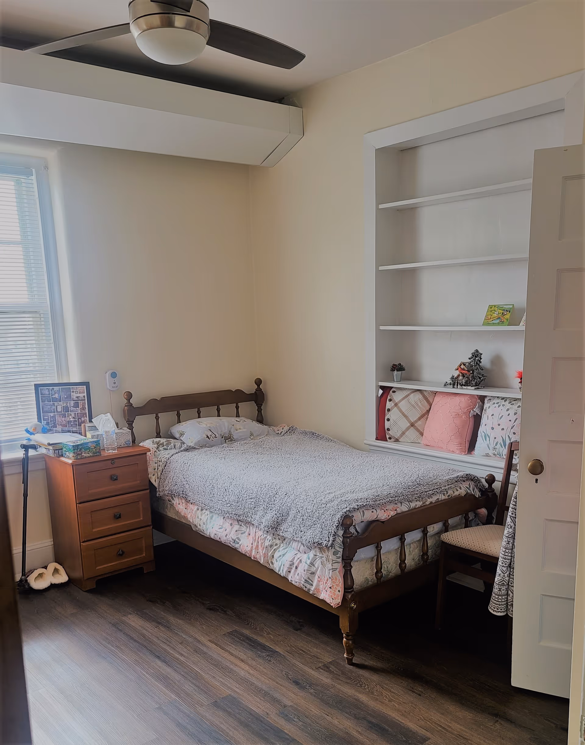 A cozy bedroom with a wooden bed frame, a bed covered with a floral bedspread and a gray blanket. Next to the bed is a wooden nightstand with three drawers, holding a framed photo collage, a tissue box, and other small items. There is a window with blinds on the left wall, a ceiling fan above, and built-in shelves on the right wall with decorative pillows and small ornaments. A wooden chair is partially visible near the door.