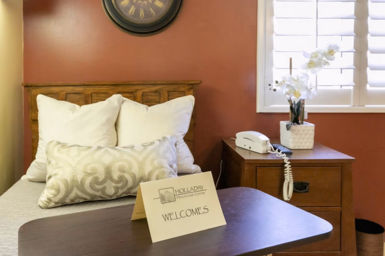 A cozy bedroom with a wooden headboard, white pillows, and a decorative cushion on the bed. A wooden nightstand beside the bed holds a white telephone and a white vase with white flowers. A clock hangs on the wall above the bed, and a window with white shutters is visible in the background. A sign on a table in front of the bed reads 'Holladay Healthcare Center WELCOMES'.