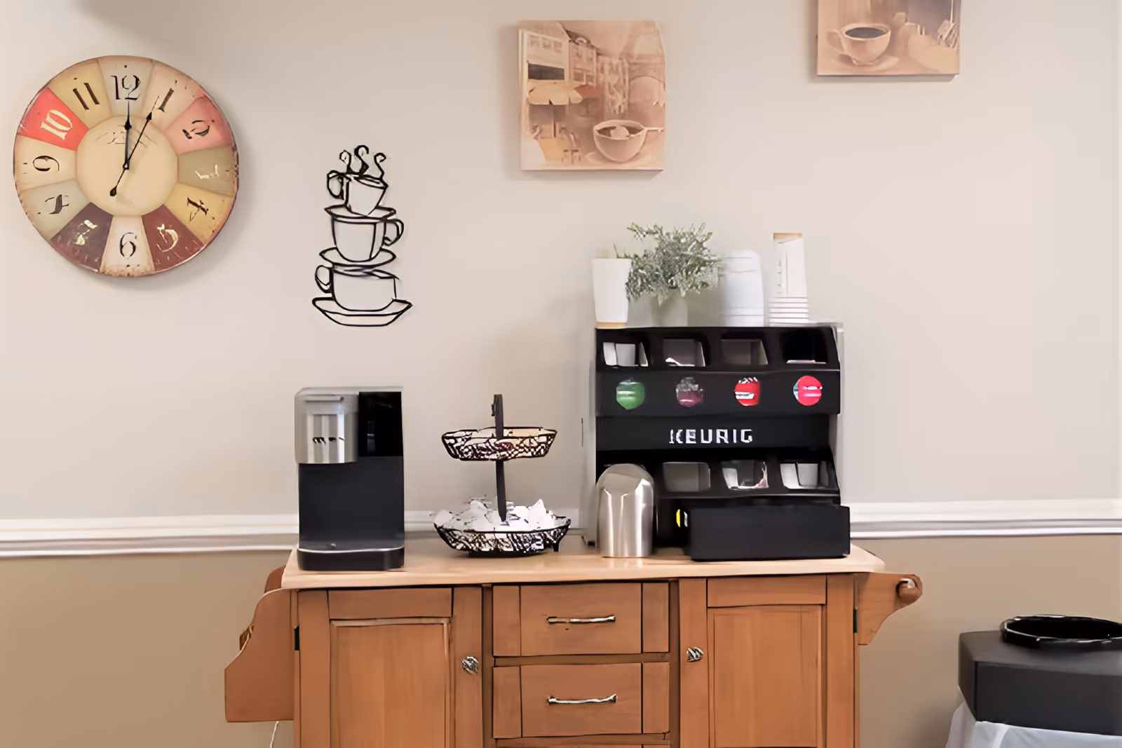 A coffee station setup on a wooden cabinet with a Keurig coffee pod organizer, a coffee machine, a two-tiered basket with coffee supplies, and a small plant in a white pot. The wall behind features a large colorful clock, a metal wall art of stacked coffee cups, and two small paintings of coffee cups.