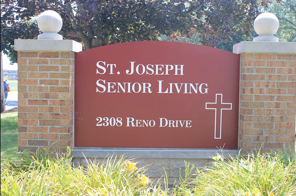 A large outdoor sign for St. Joseph Senior Living at 2308 Reno Drive, mounted between two brick pillars with decorative white spheres on top, surrounded by grass and plants with trees in the background.