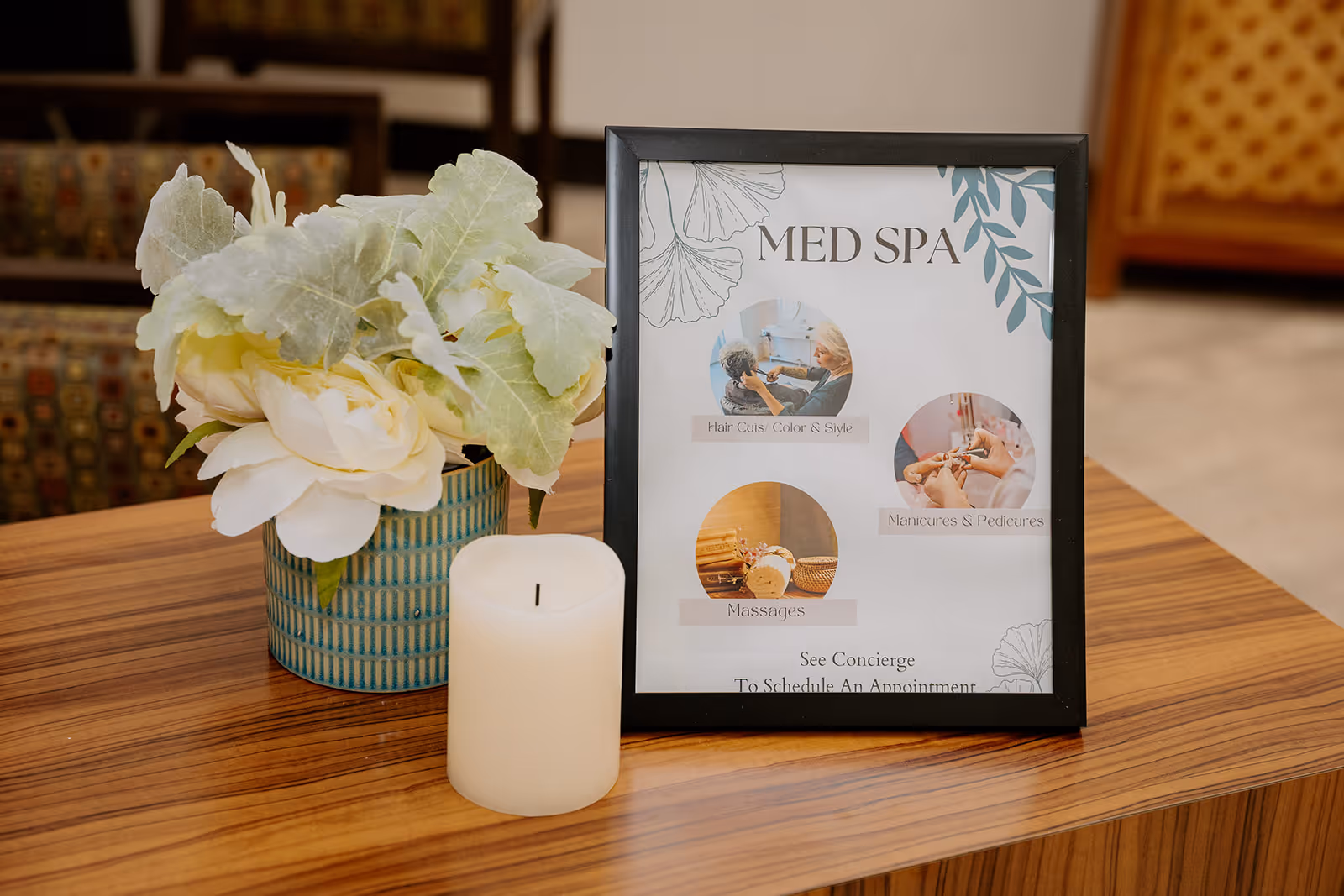 A wooden table with a small potted plant featuring white flowers and green leaves, a white candle, and a framed sign advertising Med Spa services including hair cuts, color and style, manicures and pedicures, and massages. The background shows blurred chairs and part of a room interior.