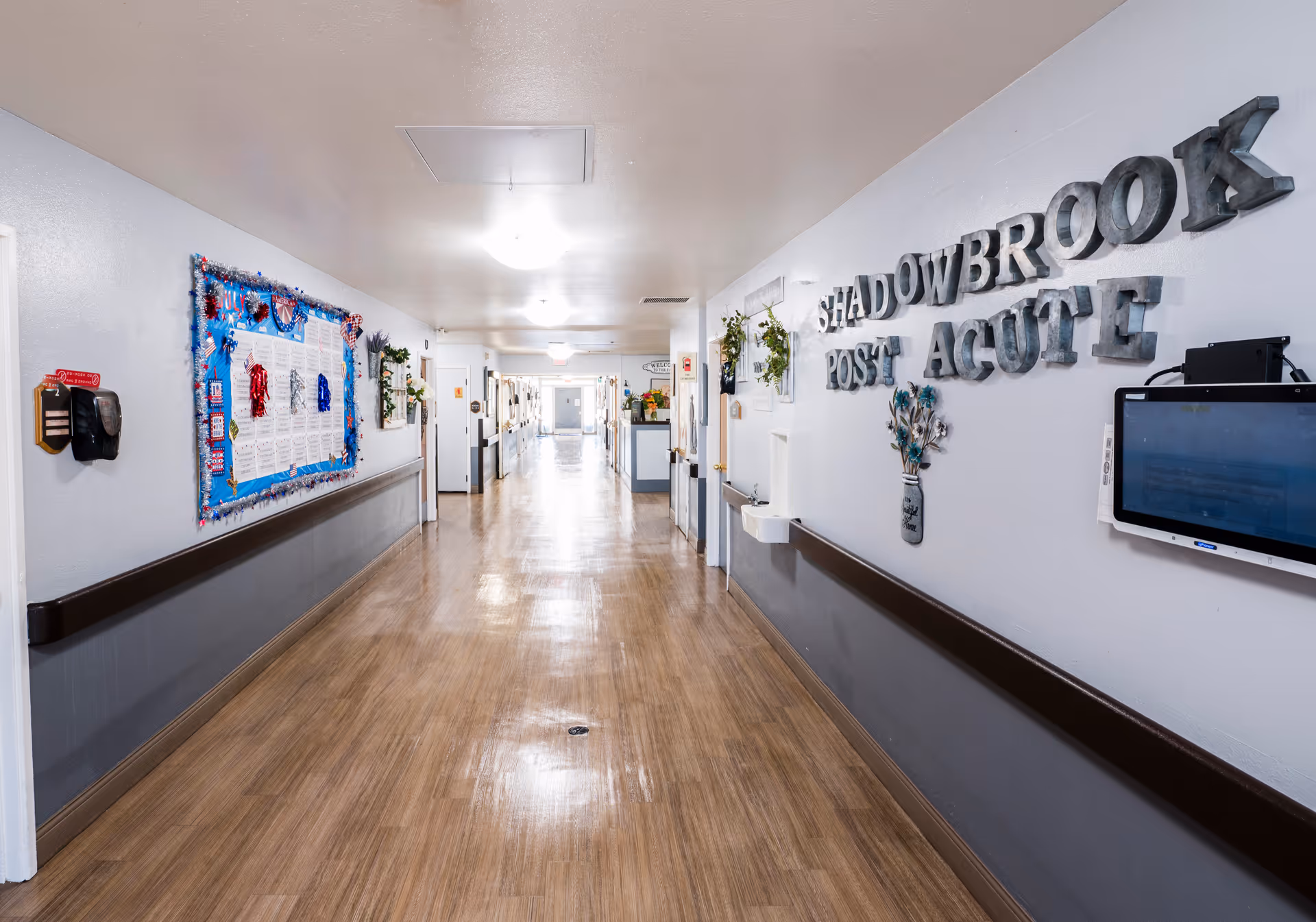 A long, clean hallway in a senior living facility with light-colored walls and wooden flooring. On the right wall, large 3D letters spell out 'SHADOWBROOK POST ACUTE' along with some decorative items and a mounted screen. The left wall features a bulletin board decorated with colorful items and notices. The hallway is well-lit with ceiling lights and has handrails on both sides.