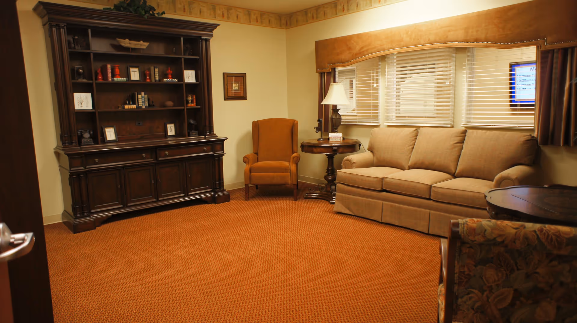 A cozy living room with a beige sofa, an orange armchair, a round wooden side table with a lamp, a large dark wooden cabinet with shelves holding decorative items, and a window with blinds and brown curtains.