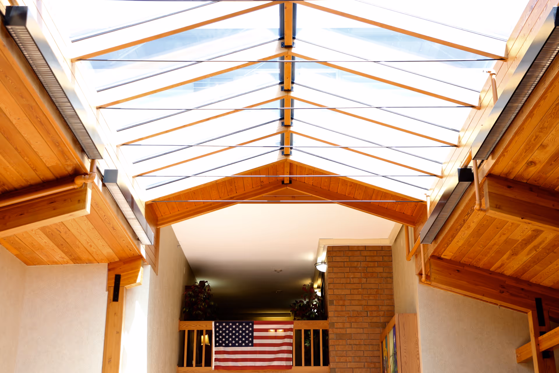 Interior view of a senior living facility with a wooden ceiling and large skylight allowing natural light to fill the space. An American flag is displayed on a balcony railing above a brick wall.