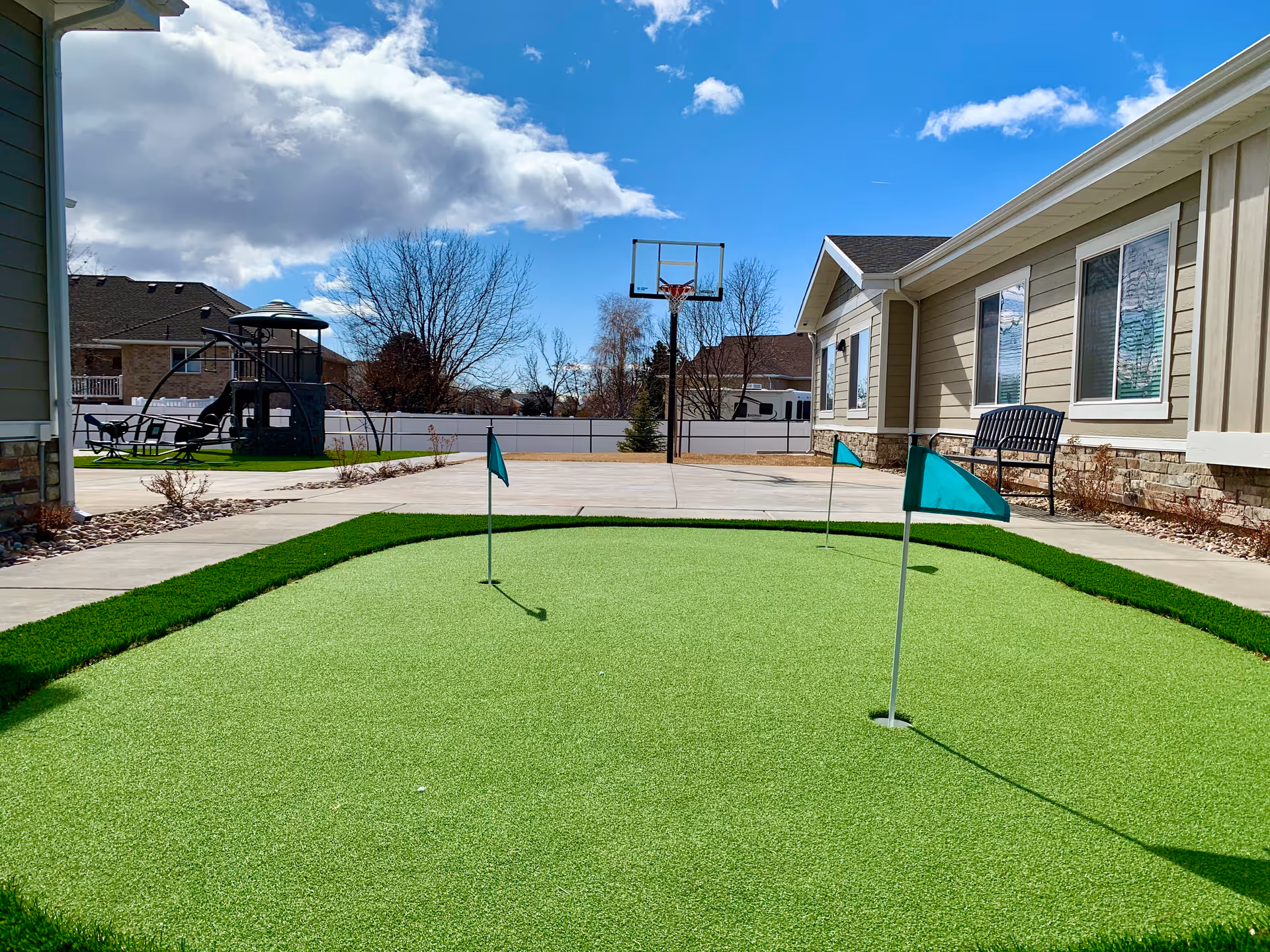 Outdoor area of Sunridge Assisted Living of Roy featuring a small putting green with three flags, a basketball hoop, a playground structure, and a bench along the side of a beige building under a partly cloudy blue sky.