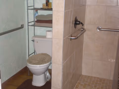 A bathroom featuring a toilet with a cushioned seat cover, a metal shelving unit above the toilet holding towels and toiletries, and a tiled walk-in shower with grab bars on the walls.