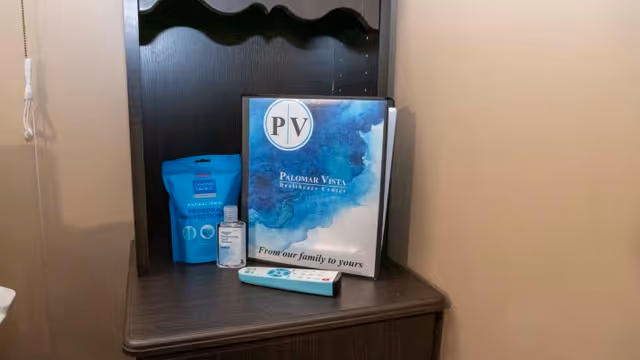 A dark wooden shelf holding a blue packet of antibacterial personal protection wipes, a small bottle of hand sanitizer, a remote control, and a binder with the Palomar Vista Healthcare Center logo and the text 'From our family to yours'.