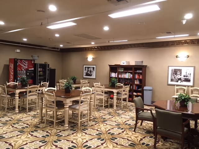 A spacious common area with several wooden tables and chairs arranged on a patterned carpet. There are plants on the tables, a bookshelf filled with books against the wall, and two black and white framed photographs hanging on the wall. In the background, there is a Coca-Cola vending machine and a refrigerator.