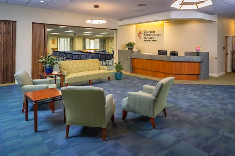A spacious reception area with a wooden front desk and a sign that reads United Methodist Homes Hilltop Campus. The room features a seating area with a green patterned sofa, three green armchairs, and a wooden coffee table. There are plants placed around the room and a large open doorway leading to a room with rows of chairs.