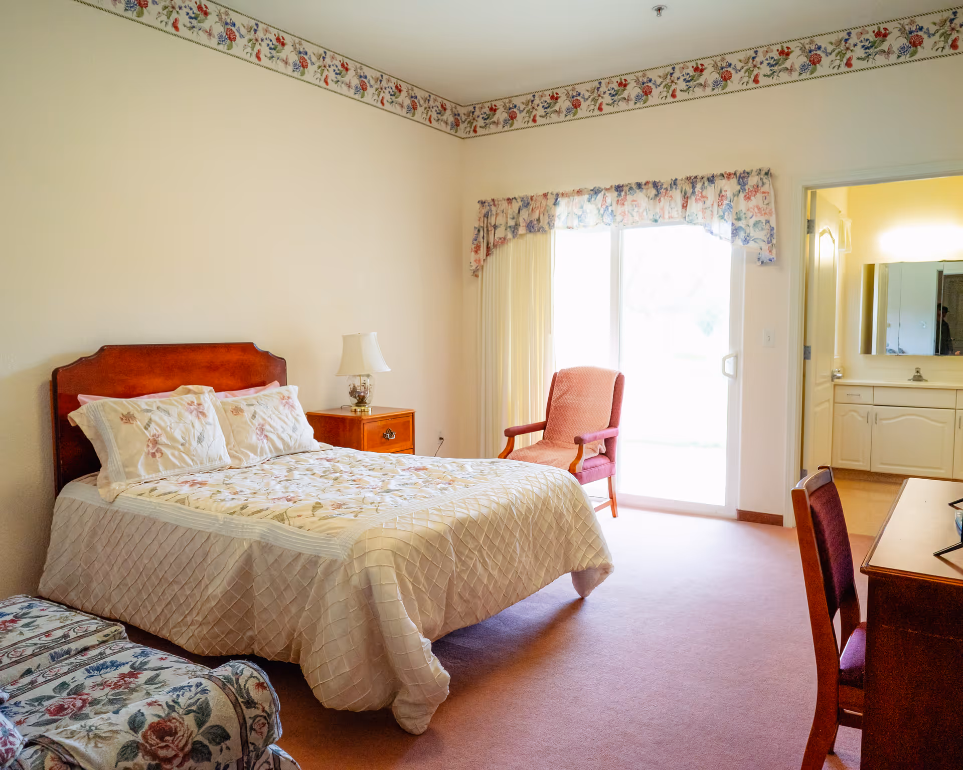 A cozy bedroom with a double bed covered in a floral quilt and matching pillows. There is a wooden nightstand with a lamp beside the bed. A pink upholstered armchair is placed near a sliding glass door with floral curtains, allowing natural light to enter the room. A wooden desk with a chair is visible on the right side, and an open doorway leads to a bathroom with a sink and mirror.