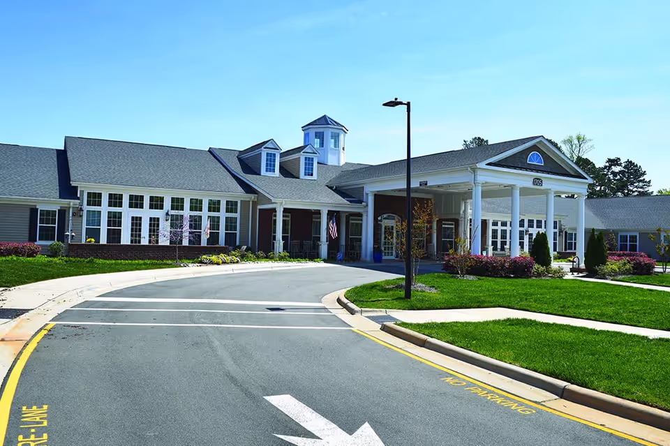 Exterior view of Spring Arbor of Cary senior living facility showing a large building with white columns at the entrance, multiple windows, a curved driveway, green lawns, and a clear blue sky.