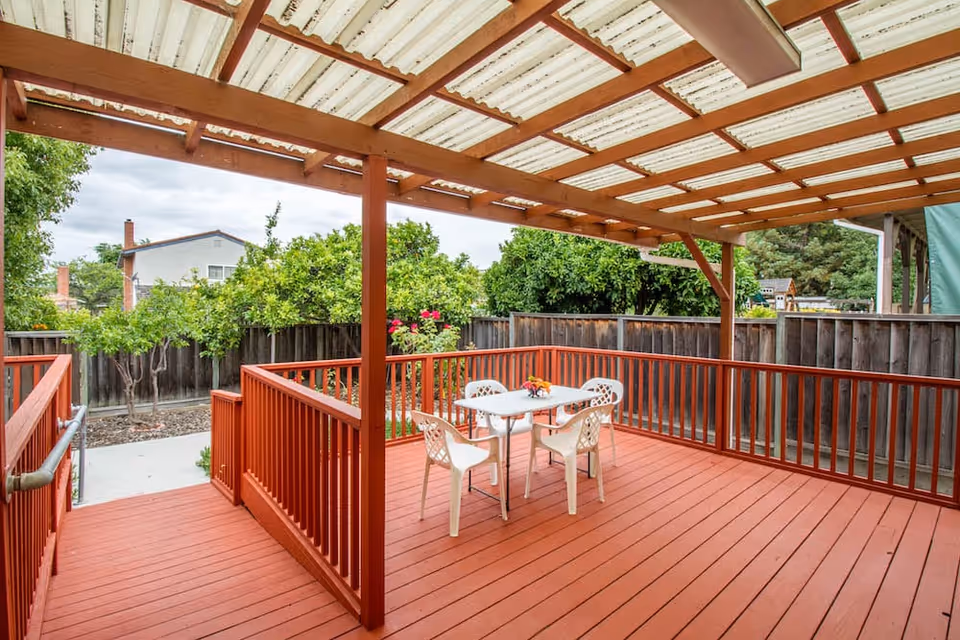 Covered red wooden deck with a small white table and four chairs overlooking a fenced backyard with trees.