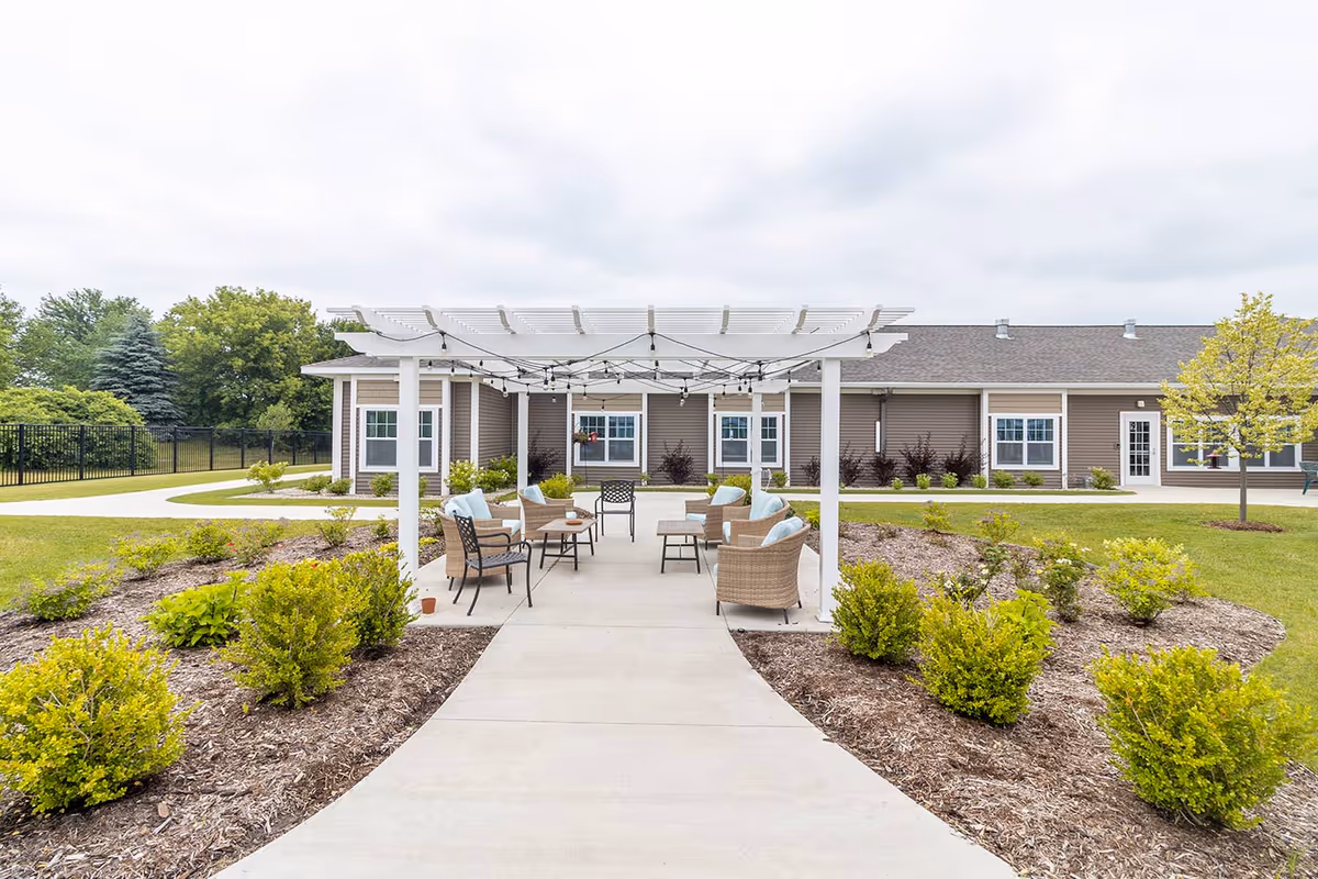 Outdoor patio with a white pergola, seating and a walkway leading to a single-story assisted living building surrounded by landscaping.