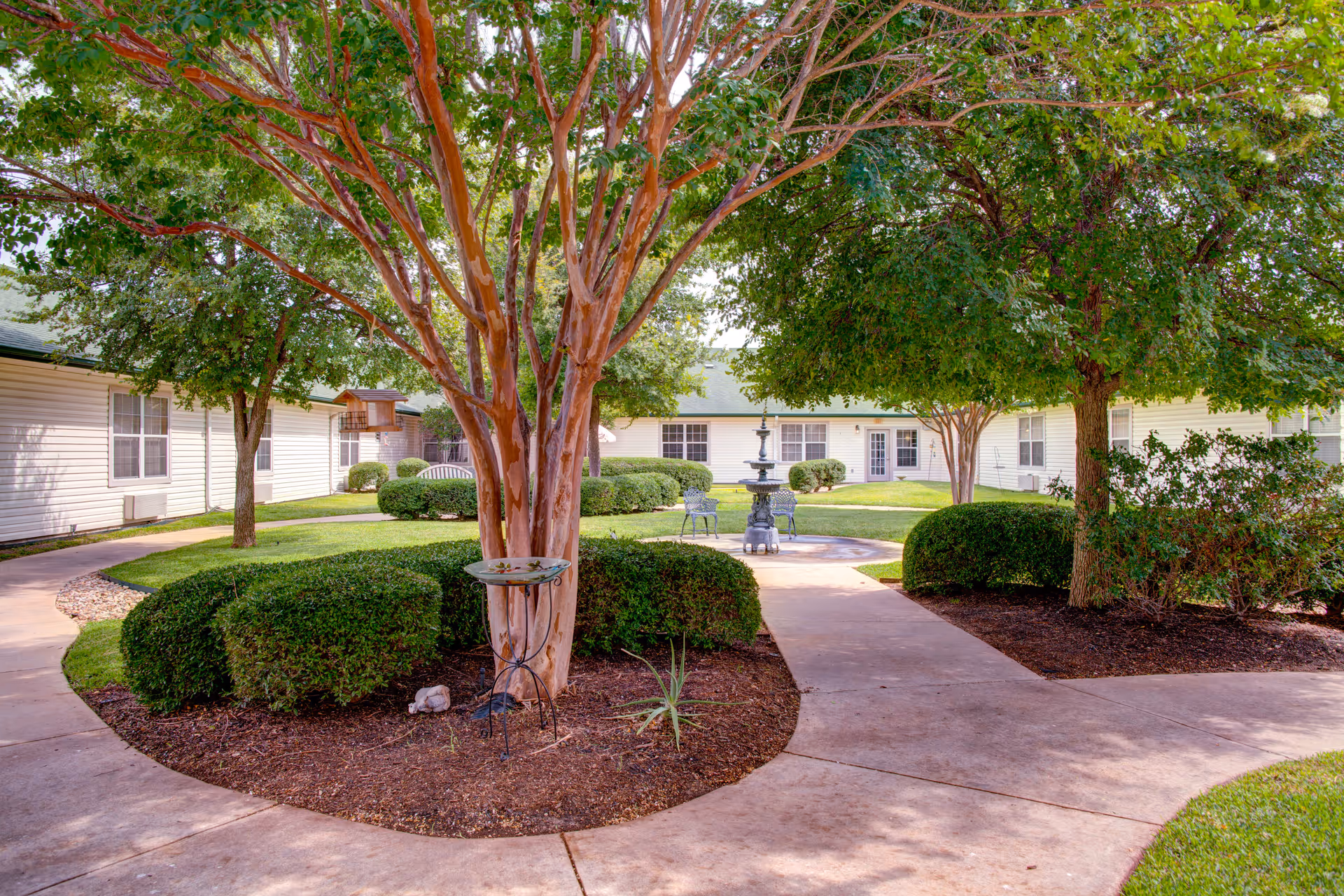 A landscaped courtyard with paved paths, mature trees, manicured shrubs, and a central fountain in front of single-story residential buildings.