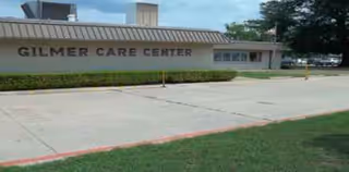 Exterior of Gilmer Care Center nursing facility with parking lot and trimmed shrubs.