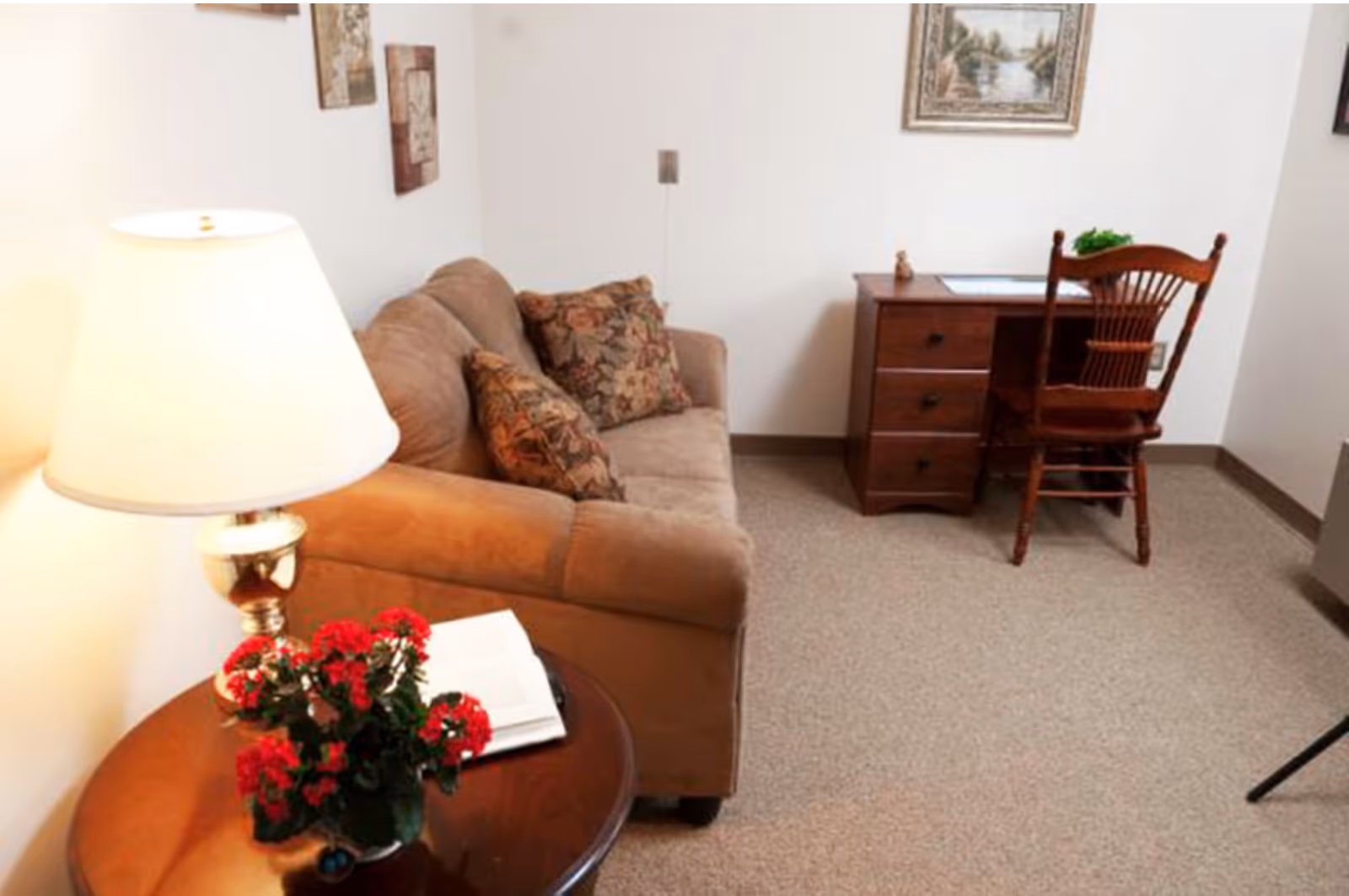 Small living area with a brown sofa, round side table holding a lamp and red flowers, and a wooden desk and chair against a white wall.