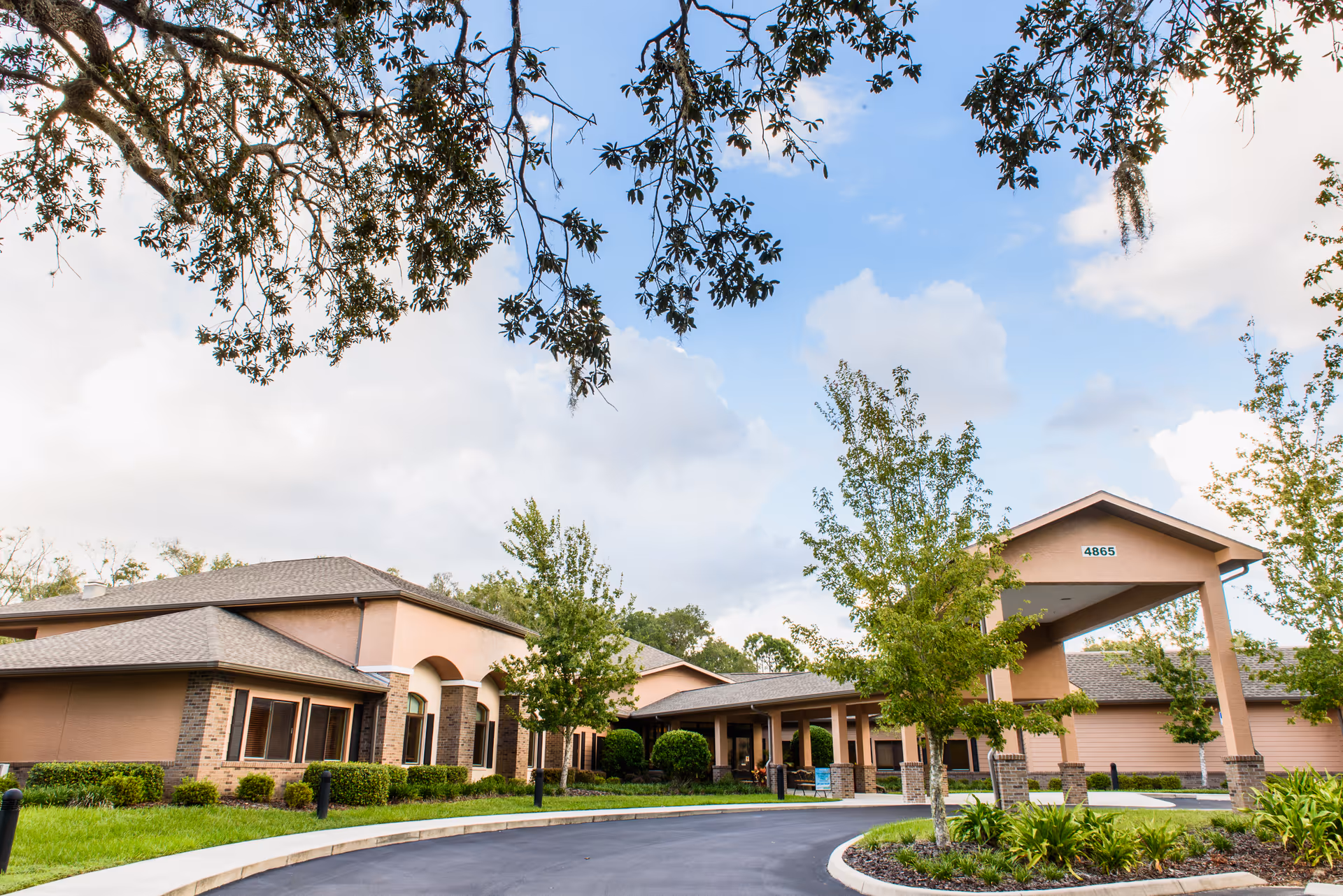 Exterior view of Best Care Senior Living at Lecanto facility showing a single-story building with a covered entrance, surrounded by trees and landscaping under a partly cloudy sky.