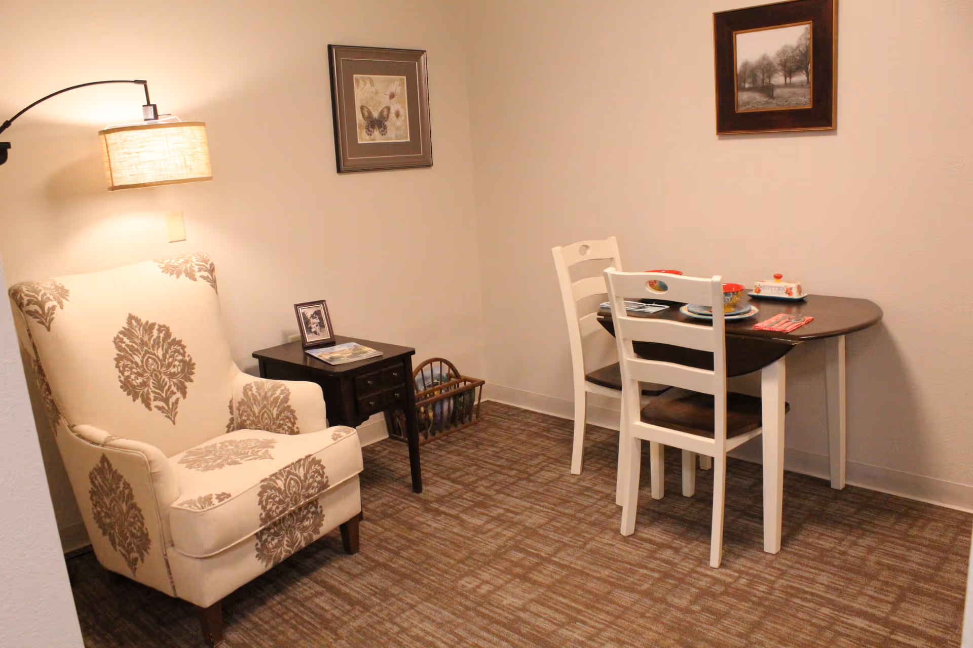 A cozy interior room with a patterned armchair and a floor lamp on the left, a small dark wooden side table with a framed photo and magazines, and a small dining table with two white chairs set with plates and utensils. The walls have two framed pictures, one of a butterfly and the other of trees.